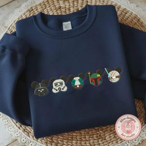 May include: Navy blue sweatshirt featuring embroidered character designs. The designs include Darth Vader, a Stormtrooper, and other characters. The sweatshirt is displayed on a woven surface.