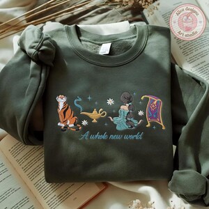 May include: A dark olive green crewneck sweatshirt featuring an embroidered graphic of a tiger, a gold oil lamp, a person with long dark hair, and a purple flying carpet. The text A whole new world is embroidered in light blue below the graphic.