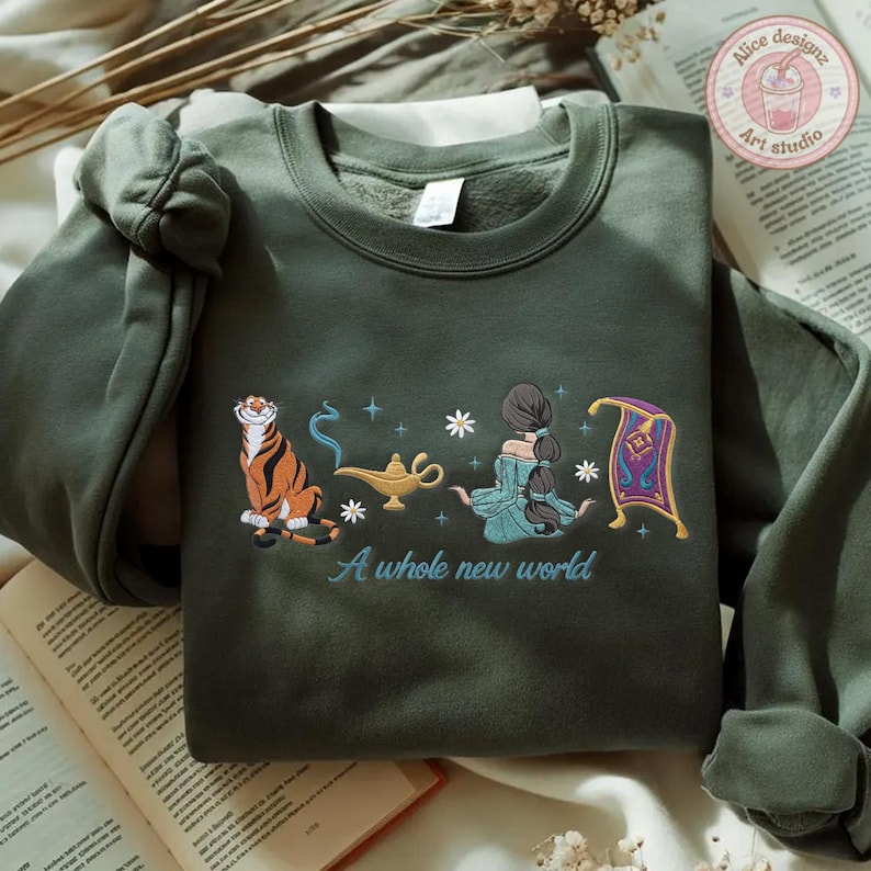 May include: A dark olive green crewneck sweatshirt featuring an embroidered graphic of a tiger, a gold oil lamp, a person with long dark hair, and a purple flying carpet. The text A whole new world is embroidered in light blue below the graphic.