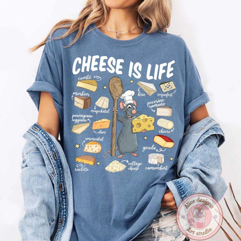 Puede incluir: Una camiseta azul con el texto "CHEESE IS LIFE" e ilustraciones de varios quesos. Una rata de dibujos animados con un gorro de chef sostiene una cuchara de madera y una cu&ntilde;a de queso. La camiseta tambi&eacute;n presenta nombres de quesos.