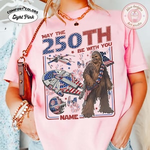 May include: Pale pink t-shirt with a Star Wars-themed graphic. The design includes the text "MAY THE 250TH BE WITH YOU" and images of the Millennium Falcon, X-wing fighters, and Chewbacca. The shirt also has the text "NAME" at the bottom.