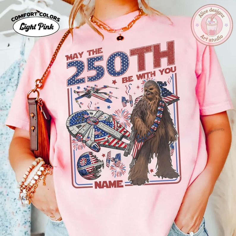 May include: Pale pink t-shirt with a Star Wars-themed graphic. The design includes the text "MAY THE 250TH BE WITH YOU" and images of the Millennium Falcon, X-wing fighters, and Chewbacca. The shirt also has the text "NAME" at the bottom.