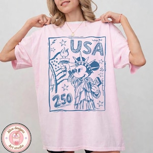May include: A light pink t-shirt featuring a blue graphic of Mickey Mouse as the Statue of Liberty, holding a torch and a book. The word "USA" is above the image, with the number "250" below. The shirt has short sleeves and a relaxed fit.