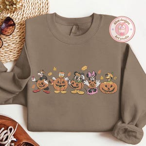 Puede incluir: Una sudadera marr&oacute;n con un dise&ntilde;o bordado de personajes de dibujos animados en calabazas talladas. El dise&ntilde;o incluye personajes como Mickey Mouse, Minnie Mouse y Donald Duck, con hojas de oto&ntilde;o. La sudadera tiene cuello redondo.