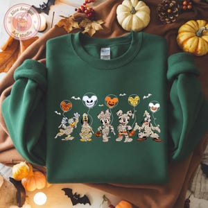 May include: A dark green sweatshirt featuring a Halloween-themed graphic. The graphic depicts cartoon characters dressed as mummies holding balloons. The balloons are shaped like Mickey Mouse, pumpkins, and ghosts. Bats are also included in the design.