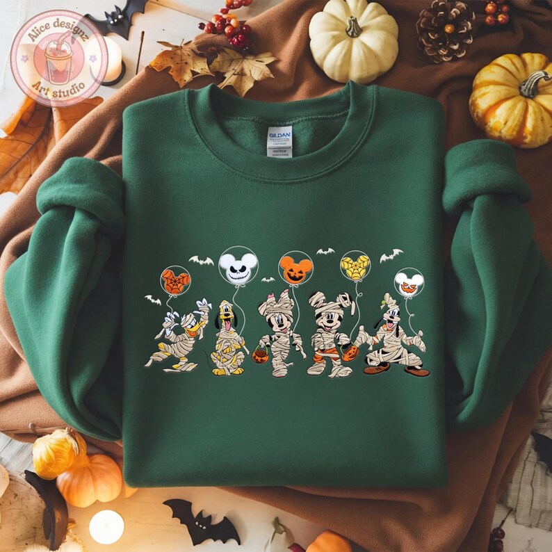 May include: A dark green sweatshirt featuring a Halloween-themed graphic. The graphic depicts cartoon characters dressed as mummies holding balloons. The balloons are shaped like Mickey Mouse, pumpkins, and ghosts. Bats are also included in the design.