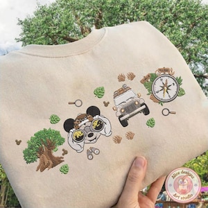 May include: Beige sweatshirt with embroidered adventure-themed graphics. Features a cartoon character with binoculars, a jeep, a compass, a tree, and paw prints. Includes green leaf and magnifying glass details. The design is in shades of brown, green, and white.