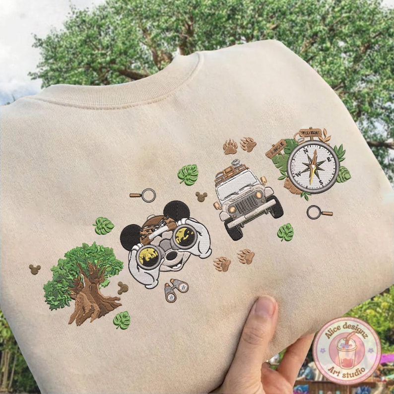 May include: Beige sweatshirt with embroidered adventure-themed graphics. Features a cartoon character with binoculars, a jeep, a compass, a tree, and paw prints. Includes green leaf and magnifying glass details. The design is in shades of brown, green, and white.