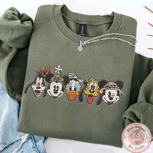 Puede incluir: Sudadera verde oliva con bordados de caras de personajes de dibujos animados, incluyendo a Goofy, Minnie Mouse, Donald Duck, Pluto y Mickey Mouse. Un collar plateado con un colgante est&aacute; sobre la sudadera.