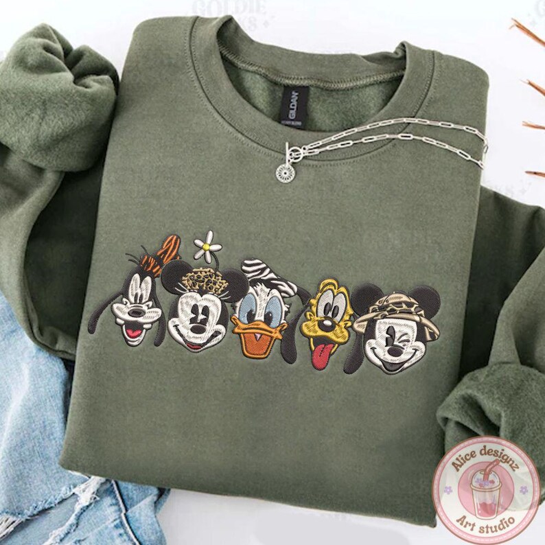 Puede incluir: Sudadera verde oliva con bordados de caras de personajes de dibujos animados, incluyendo a Goofy, Minnie Mouse, Donald Duck, Pluto y Mickey Mouse. Un collar plateado con un colgante est&aacute; sobre la sudadera.