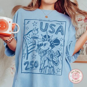 May include: Light blue t-shirt featuring a sketch-style graphic of Mickey Mouse as the Statue of Liberty, holding a torch and a book that says "July". The word "USA" is above the character. The number "250" is below the character. The shirt has a relaxed fit.