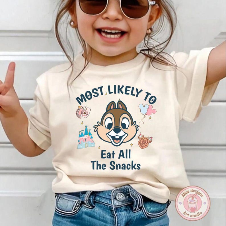 May include: Beige t-shirt with the text "MOST LIKELY TO Eat All The Snacks" and a cartoon graphic of Chip from Chip 'n Dale. The graphic includes a camera, balloons, a castle, and a cookie. The shirt is worn by a child.