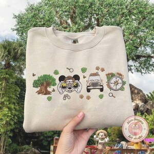 May include: Beige sweatshirt with embroidered safari-themed design. Features a tree, binoculars, a jeep, and a compass. The design includes Mickey Mouse ears and paw prints. The sweatshirt is held up by a person.