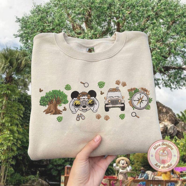 May include: Beige sweatshirt with embroidered safari-themed design. Features a tree, binoculars, a jeep, and a compass. The design includes Mickey Mouse ears and paw prints. The sweatshirt is held up by a person.