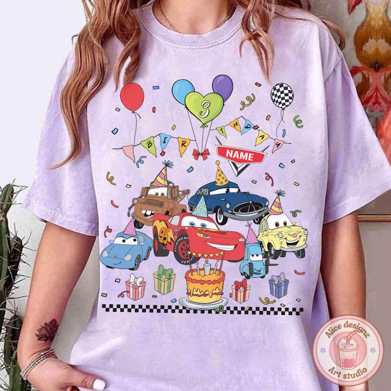 Op de afbeelding: Lichtpaarse t-shirt met een kleurrijke afbeelding van tekenfilmauto's die een verjaardag vieren. Het ontwerp bevat een verjaardagstaart, ballonnen, confetti en een banner met het woord "NAME". Het nummer "3" is ook aanwezig.