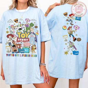 May include: A light blue t-shirt featuring a colorful graphic print of Toy Story characters on the front and back. The front displays the Toy Story logo with the text You've Got A Friend In Me. The back features various characters and icons.