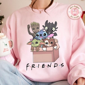Peut inclure: Un sweat-shirt rose clair avec un dessin anim&eacute; de personnages anim&eacute;s dans une bo&icirc;te. Le mot "FRIENDS" est imprim&eacute; en noir en bas. Les personnages incluent Baby Yoda, Stitch, Groot, un Night Fury et un Mogwai.