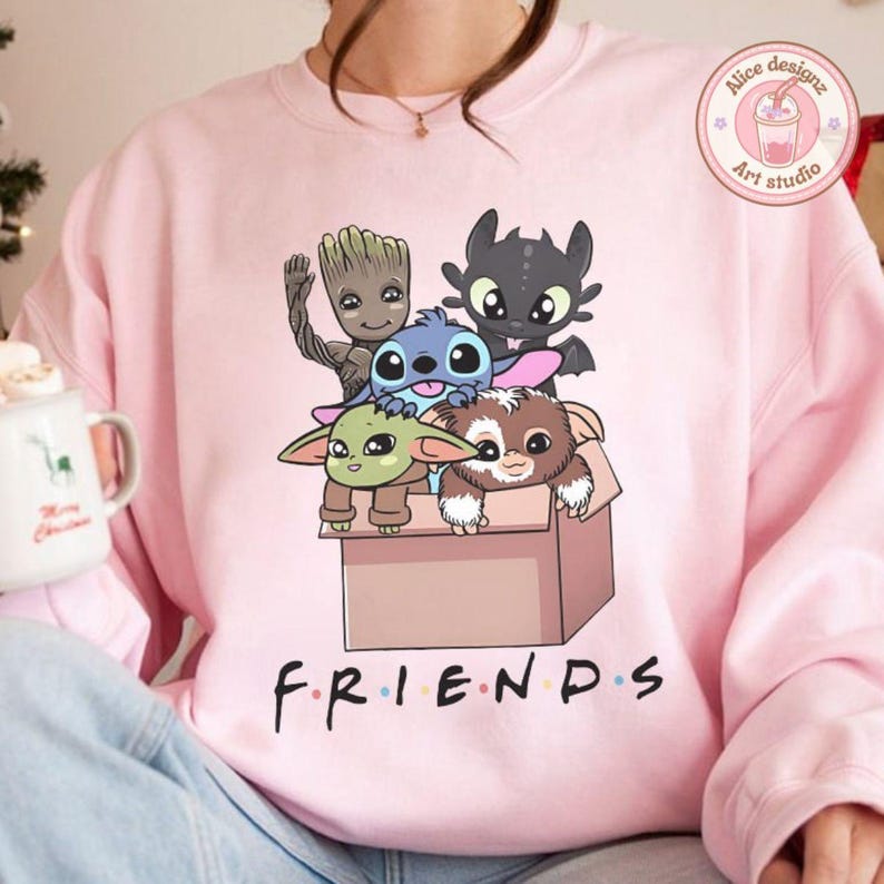 Peut inclure: Un sweat-shirt rose clair avec un dessin anim&eacute; de personnages anim&eacute;s dans une bo&icirc;te. Le mot "FRIENDS" est imprim&eacute; en noir en bas. Les personnages incluent Baby Yoda, Stitch, Groot, un Night Fury et un Mogwai.