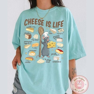 Puede incluir: Camiseta azul claro con la frase "CHEESE IS LIFE" e ilustraciones de varios quesos. Un chef de dibujos animados con forma de rata sostiene una cuchara de madera y una cu&ntilde;a de queso. La camiseta tambi&eacute;n presenta el texto "Alice designz Art studio".