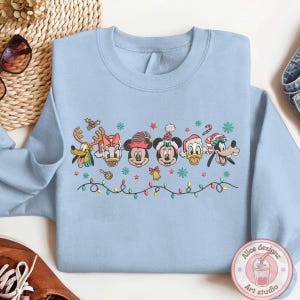 Op de afbeelding: Lichtblauw sweatshirt met geborduurde Disney-personages in kerstoutfits. Het ontwerp bevat Mickey Mouse, Minnie Mouse, Donald Duck, Katrien Duck, Goofy en Pluto, plus feestelijke elementen zoals sneeuwvlokken en een lichtsnoer.