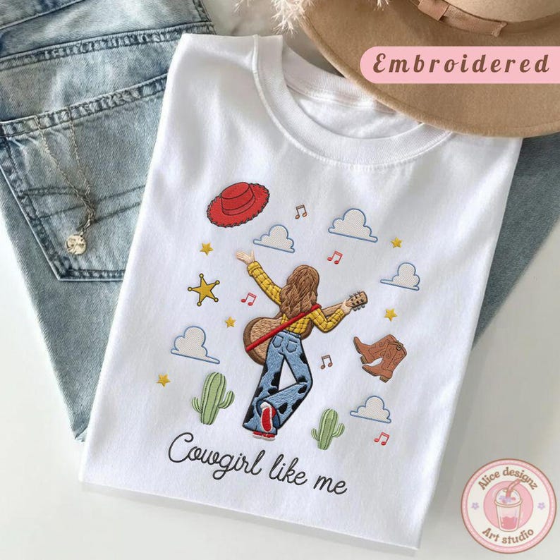 May include: A white t-shirt featuring an embroidered design of a person with long brown hair playing an acoustic guitar. The graphic includes a red cowboy hat, blue cow-print pants, brown boots, cacti, clouds, stars, and musical notes. The text reads Cowgirl like me.