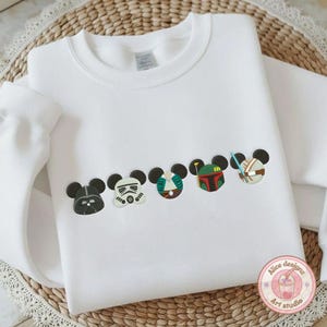 May include: White sweatshirt featuring embroidered Star Wars-themed Mickey Mouse heads. The design includes Darth Vader, a Stormtrooper, and other characters. The sweatshirt is made of a soft material and is suitable for casual wear.