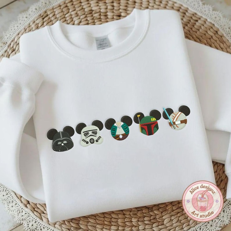 May include: White sweatshirt featuring embroidered Star Wars-themed Mickey Mouse heads. The design includes Darth Vader, a Stormtrooper, and other characters. The sweatshirt is made of a soft material and is suitable for casual wear.