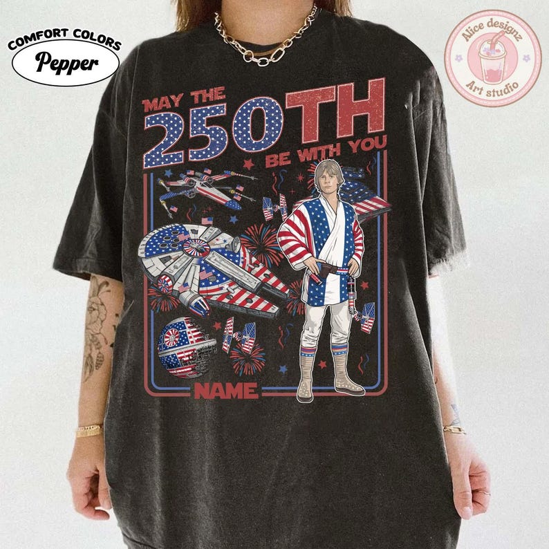 May include: Dark grey t-shirt with a patriotic design. The graphic includes the text "MAY THE 250TH BE WITH YOU" with a Star Wars theme, featuring a character in a patriotic outfit, a Millennium Falcon, and fireworks.