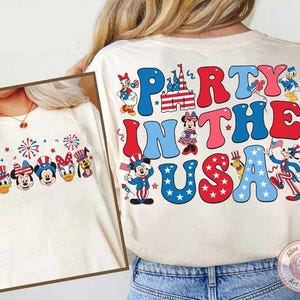 Peut inclure: T-shirt cr&egrave;me avec un motif patriotique portant l'inscription "Party in the USA". Le motif comprend des personnages de dessins anim&eacute;s comme Mickey Mouse, Minnie Mouse, Donald Duck et Dingo, ainsi que des feux d'artifice et le drapeau am&eacute;ricain.