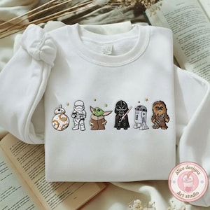 May include: White sweatshirt featuring embroidered Star Wars characters: BB-8, a Stormtrooper, Yoda, Darth Vader, R2-D2, and Chewbacca. The design includes small star accents. The sweatshirt has long sleeves and a crew neck.