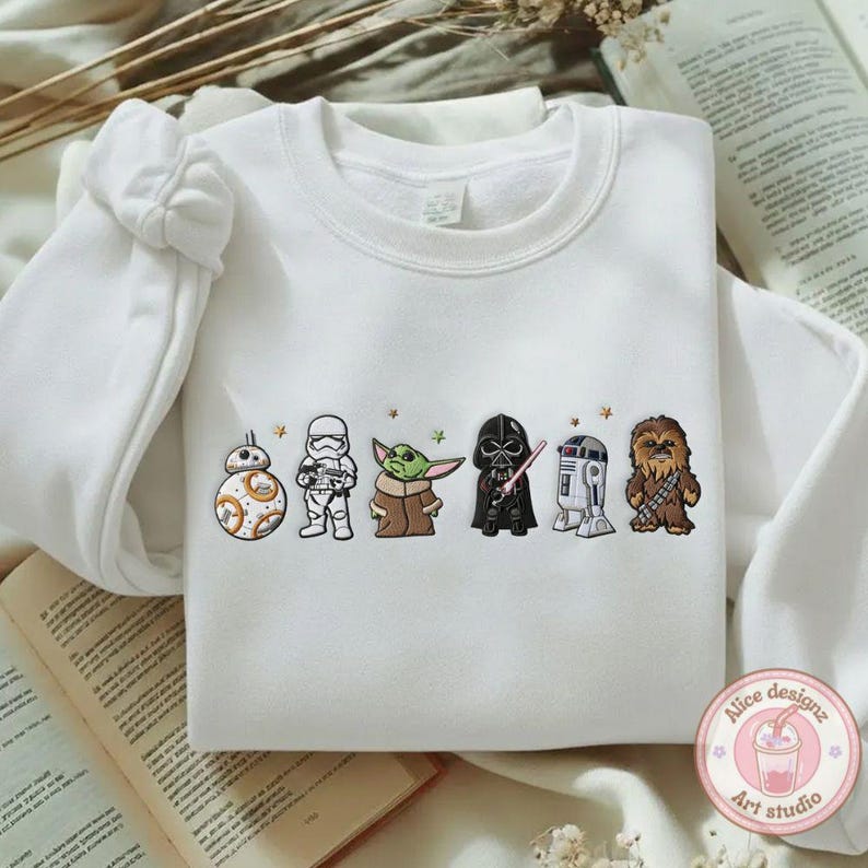 May include: White sweatshirt featuring embroidered Star Wars characters: BB-8, a Stormtrooper, Yoda, Darth Vader, R2-D2, and Chewbacca. The design includes small star accents. The sweatshirt has long sleeves and a crew neck.