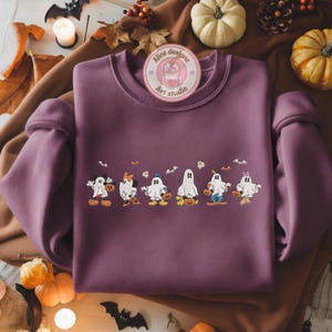 K&ouml;nnte beinhalten: Ein mauvefarbenes Sweatshirt mit Rundhalsausschnitt und einer Reihe von Cartoon-Geistern, die als beliebte Charaktere verkleidet sind und jeweils einen K&uuml;rbis halten. Das Design umfasst Flederm&auml;use und K&uuml;rbisse. Das Sweatshirt hat den Text "Alice designz Art studio".