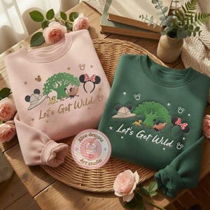 May include: Two crewneck sweatshirts, one pink and one green, with a cartoon graphic. The graphic includes a tree, safari hat, Minnie Mouse ears, and the text "Let's Get Wild". A patch with a drink design is also visible.