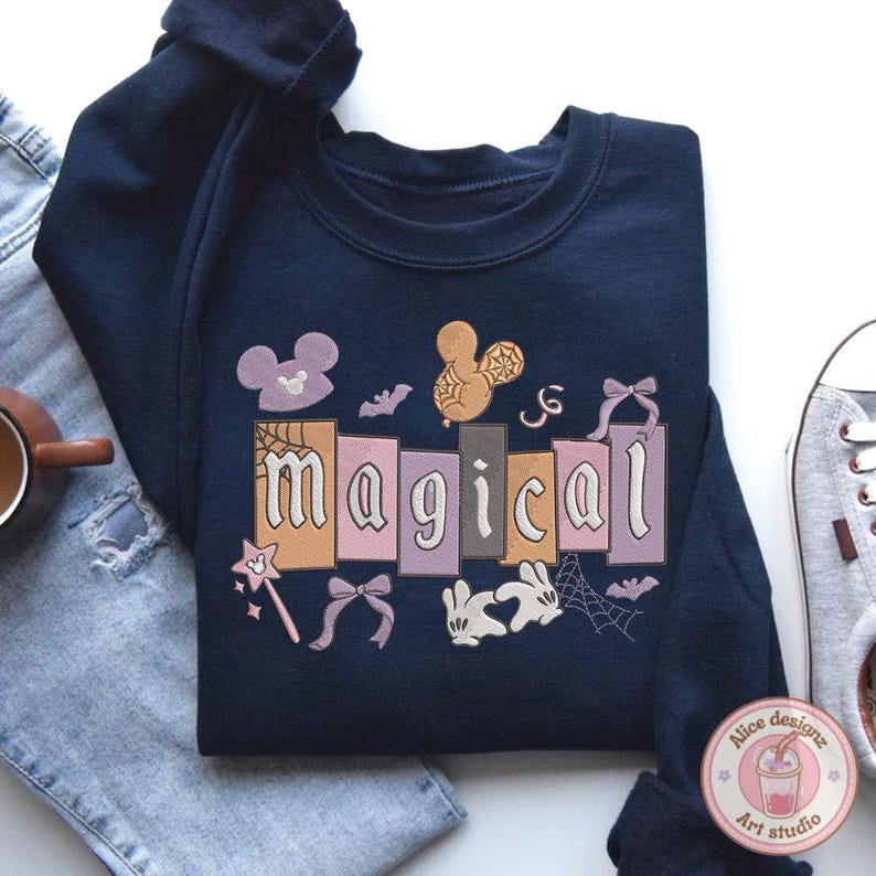 May include: A navy blue sweatshirt featuring the word "magical" in a decorative font, with cartoon illustrations of bats, bows, and Mickey Mouse-style shapes. The sweatshirt has a crew neck and long sleeves. Jeans and a trainer are in the background.