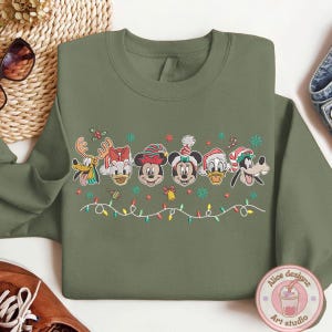Op de afbeelding: Olijfgroene sweatshirt met geborduurde Disney-figuren in feestelijke kerstoutfits. Het ontwerp bevat Mickey, Minnie, Donald, Daisy, Goofy en Pluto, met kerstverlichting en sneeuwvlokken.