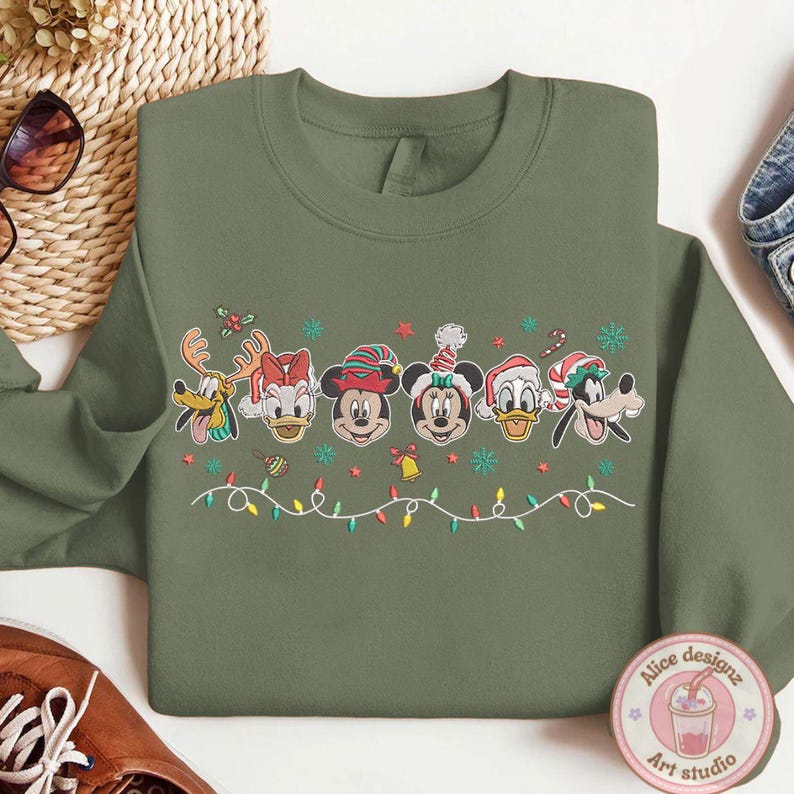 Op de afbeelding: Olijfgroene sweatshirt met geborduurde Disney-figuren in feestelijke kerstoutfits. Het ontwerp bevat Mickey, Minnie, Donald, Daisy, Goofy en Pluto, met kerstverlichting en sneeuwvlokken.