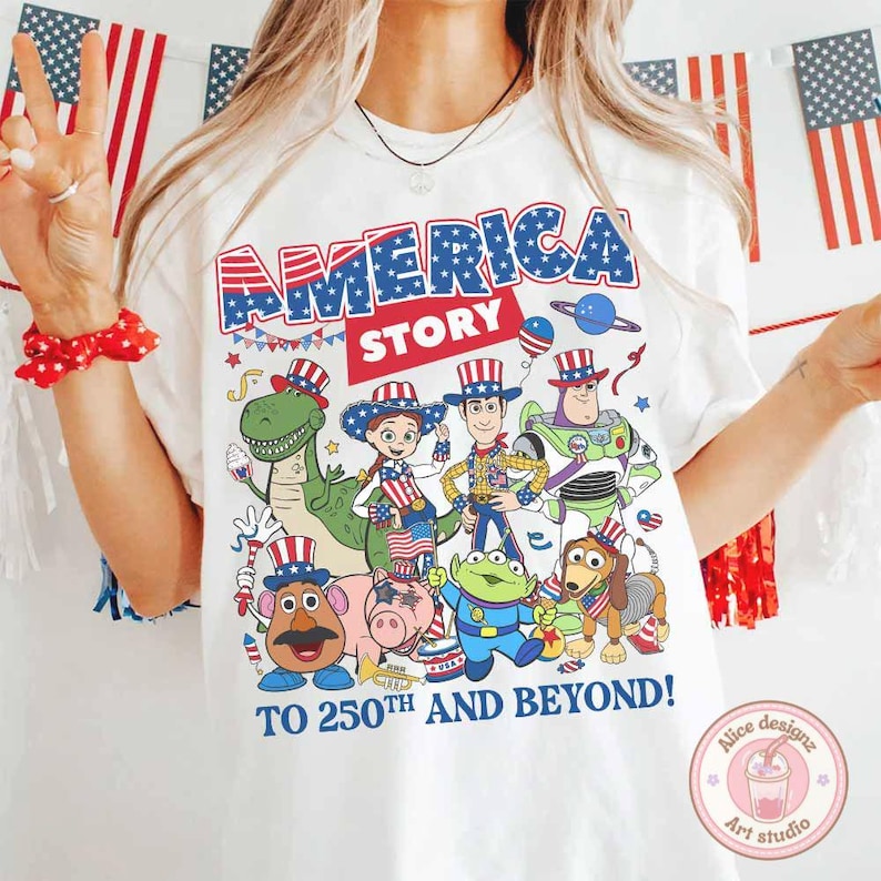 May include: White t-shirt with the text "AMERICA STORY" in blue and red, featuring cartoon characters in patriotic attire. The phrase "TO 250TH AND BEYOND!" is printed below the characters. The shirt has a round neck.