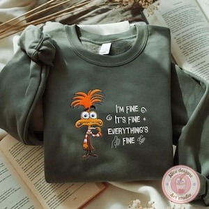 May include: An olive green sweatshirt featuring an embroidered cartoon character with orange hair and a striped scarf. The text "I'M FINE, IT'S FINE, EVERYTHING'S FINE" is embroidered in white. The sweatshirt has a crew neck and long sleeves.