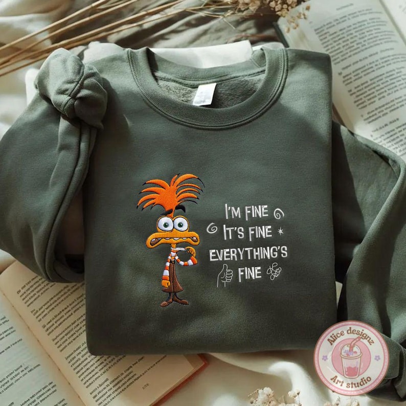May include: An olive green sweatshirt featuring an embroidered cartoon character with orange hair and a striped scarf. The text "I'M FINE, IT'S FINE, EVERYTHING'S FINE" is embroidered in white. The sweatshirt has a crew neck and long sleeves.