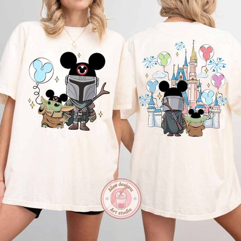 May include: Cream-colored t-shirt featuring a cartoon illustration of The Mandalorian, Grogu, and a Mickey Mouse balloon on the front. The back of the shirt shows the characters in front of a Disney castle with balloons and fireworks.