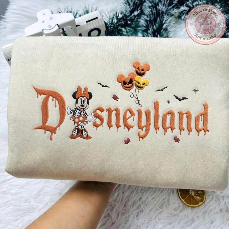May include: A cream-colored sweatshirt featuring an embroidered design with the word Disneyland in an orange dripping font. The design includes a skeleton Minnie Mouse, three jack-o'-lantern balloons, and small black bats and stars.
