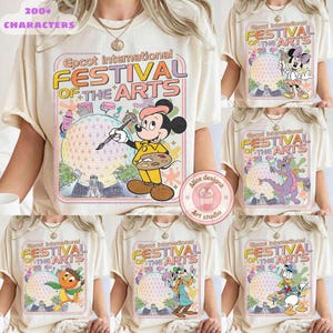 May include: Cream-colored t-shirts featuring the "Epcot International Festival of the Arts" logo with various Disney characters. Mickey Mouse, Minnie Mouse, Figment, Orange Bird, Goofy, and Donald Duck are depicted with art supplies. The shirts have a vintage aesthetic.