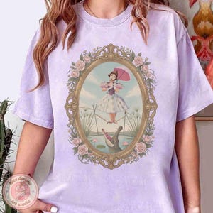 May include: A light purple t-shirt featuring a central graphic inside an ornate gold oval frame. The illustration depicts a person in a dress holding a pink umbrella, balancing on a tightrope above a green alligator in a pond with pink flowers.