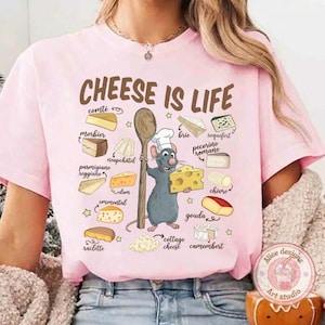 Puede incluir: Camiseta rosa claro con la frase "CHEESE IS LIFE" e ilustraciones de quesos. Un chef de dibujos animados, una rata, sostiene un trozo de queso y una cuchara de madera. Quesos: Brie, Gouda, Parmesano.