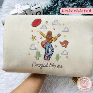 May include: A cream-colored sweatshirt featuring an embroidered design of a person with long brown hair playing a guitar, wearing a yellow shirt and cow-print pants. The design includes a red hat, brown boots, cacti, clouds, stars, and musical notes, with the text "Cowgirl like me" below.