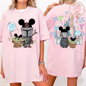 May include: Light pink t-shirt featuring a cartoon illustration of The Mandalorian and Grogu with Mickey Mouse ears. The front design includes a blue balloon. The back design shows a castle and fireworks.