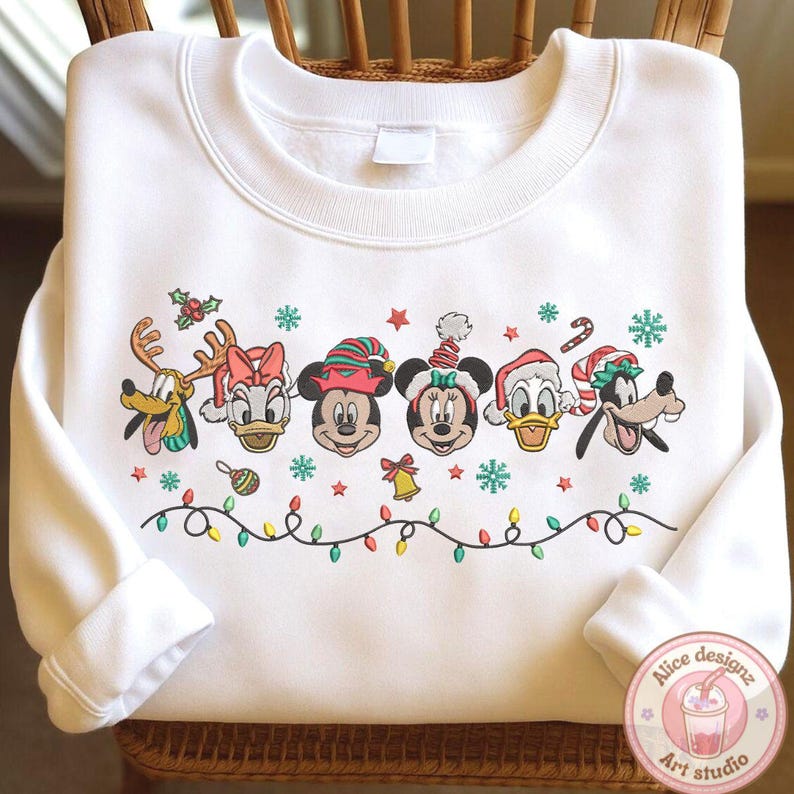Op de afbeelding: Witte sweatshirt met geborduurde Disney-personages in kerstoutfits. Het ontwerp bevat Mickey en Minnie Mouse, Donald Duck, Goofy en anderen, met sneeuwvlokken, sterren en een lichtslinger. De sweatshirt is op een houten stoel gevouwen.