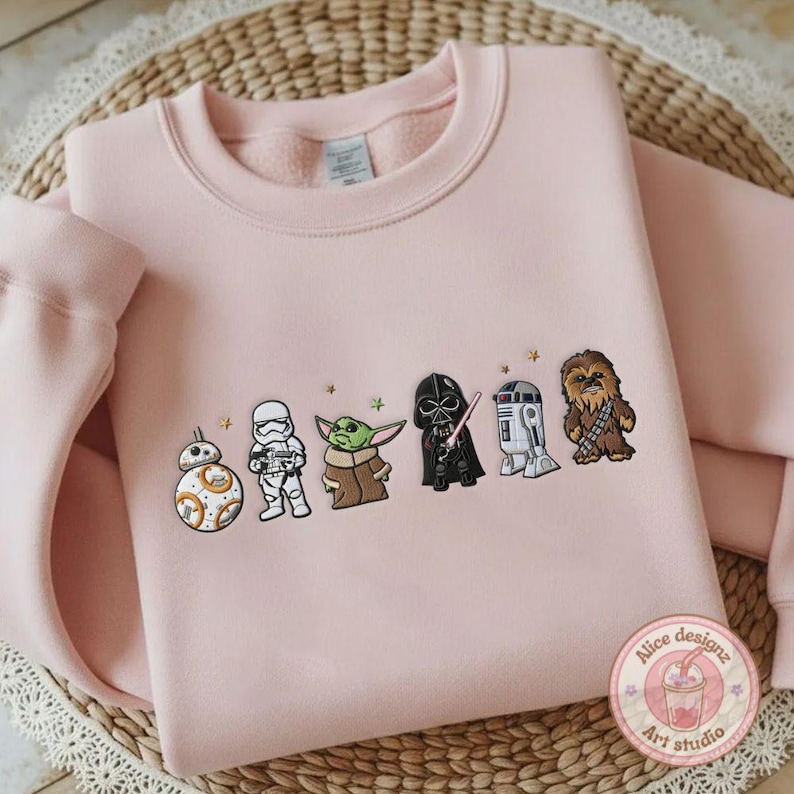 May include: A light pink sweatshirt featuring embroidered Star Wars characters: BB-8, a Stormtrooper, Grogu, Darth Vader, R2-D2, and Chewbacca. The design includes small star accents. The sweatshirt is folded on a woven surface.