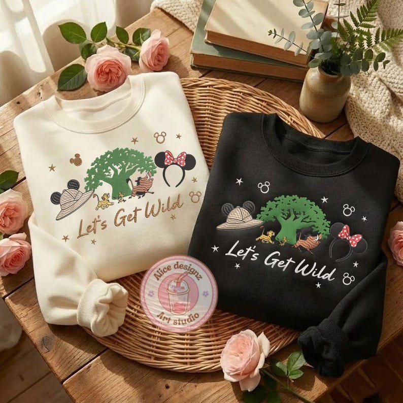 May include: Two sweatshirts, one cream and one black, with a "Let's Get Wild" design. The design includes a tree, safari hat, Minnie Mouse ears, and cartoon characters. A round sticker with text is visible.