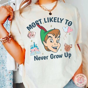 May include: Cream-colored t-shirt featuring Peter Pan with the text "Most Likely To Never Grow Up." The design includes a pink camera, a castle, balloons, and a Mickey Mouse-shaped treat. The shirt has a round neck and short sleeves.